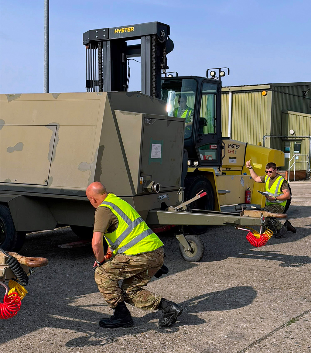 Personnel from No. 4624 Movements Squadron, Royal Auxiliary Air Force, use a Hyster heavy-duty forklift to lift and position a large tan generator trailer outside a military building.